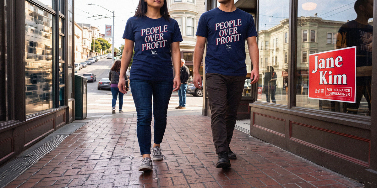 Two people walking down a city sidewalk wearing matching navy blue t-shirts with the slogan 'PEOPLE OVER PROFIT' printed in white. A red sign on the storefront reads 'Jane Kim'.